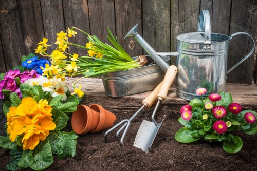 A garden scene featuring a freshly turned flower bed with dark, rich soil in the foreground, surrounded by vibrant blooming flowers in shades of yellow, purple, white, and pink. To the left, there are potted plants with large green leaves, while on the right, a cluster of pink and yellow primroses adds a splash of colour. Behind the flowers, gardening tools including a trowel, a hand fork, and a watering can are arranged on a wooden garden fence, which provides a rustic backdrop. The scene suggests active gardening work, with a focus on flower planting and bed preparation typical in a well-maintained outdoor space in Notting Hill, London, capturing a moment of gardening activity suitable for showcasing comprehensive gardening services by Garden Maintenance Notting Hill.